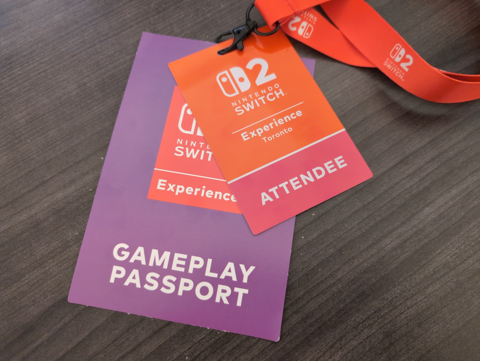 Photo of the lanyard, attendee badge, and Gameplay Passport from the event laying on a desk. They all have simple designs with big white Nintendo Switch 2 logos on red backgrounds, though the Gameplay Passport is otherwise purple.