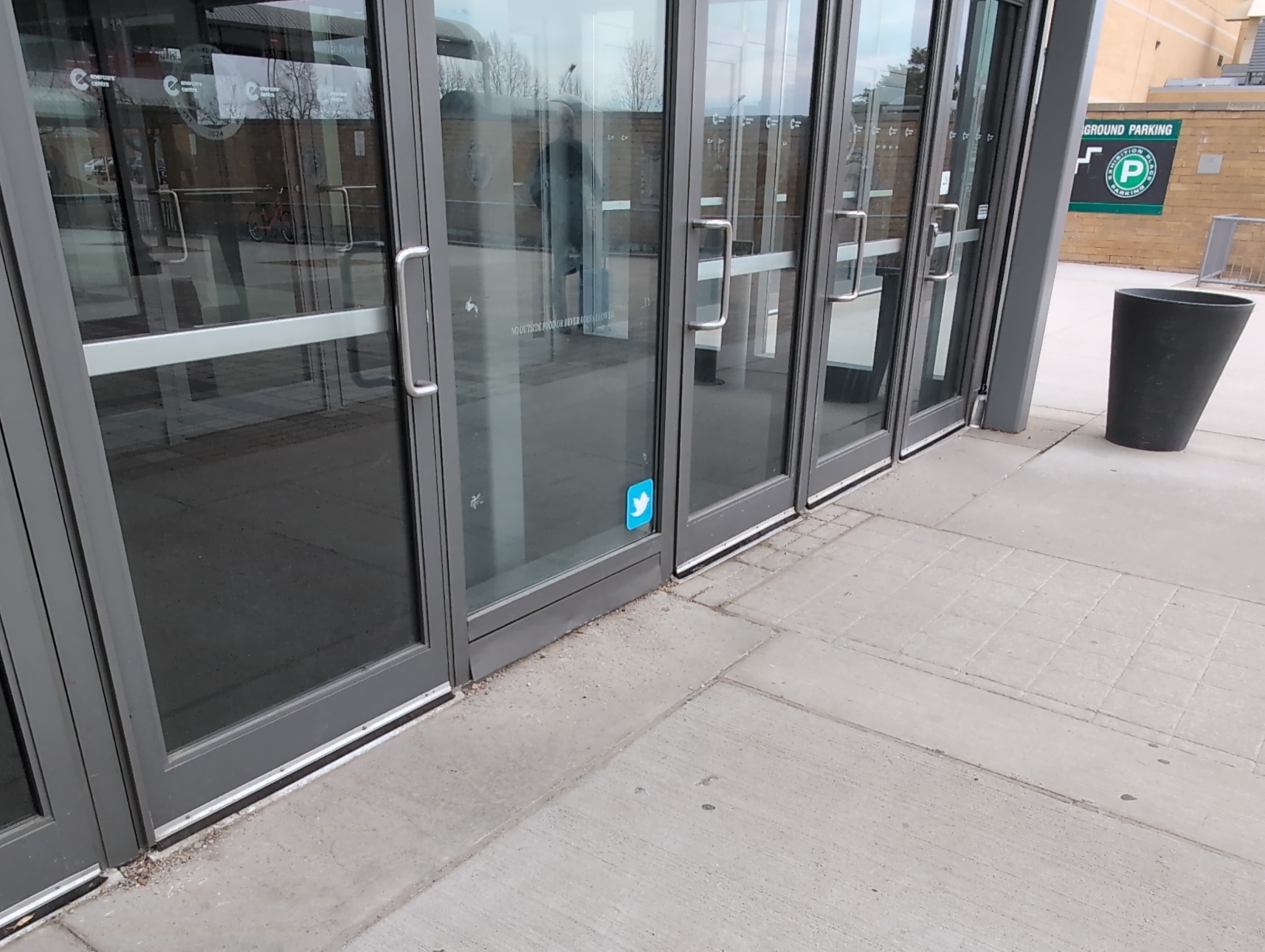 Photo of the glass building entrance. In the very centre of the frame is the bottom‐right corner of a large pane of glass separating the 2 sets of doors, which has a rounded blue sticker just above ground level containing nothing but a white Twitter logo, specifically the first bird icon used from 2010–2012. No other stickers are present anywhere.