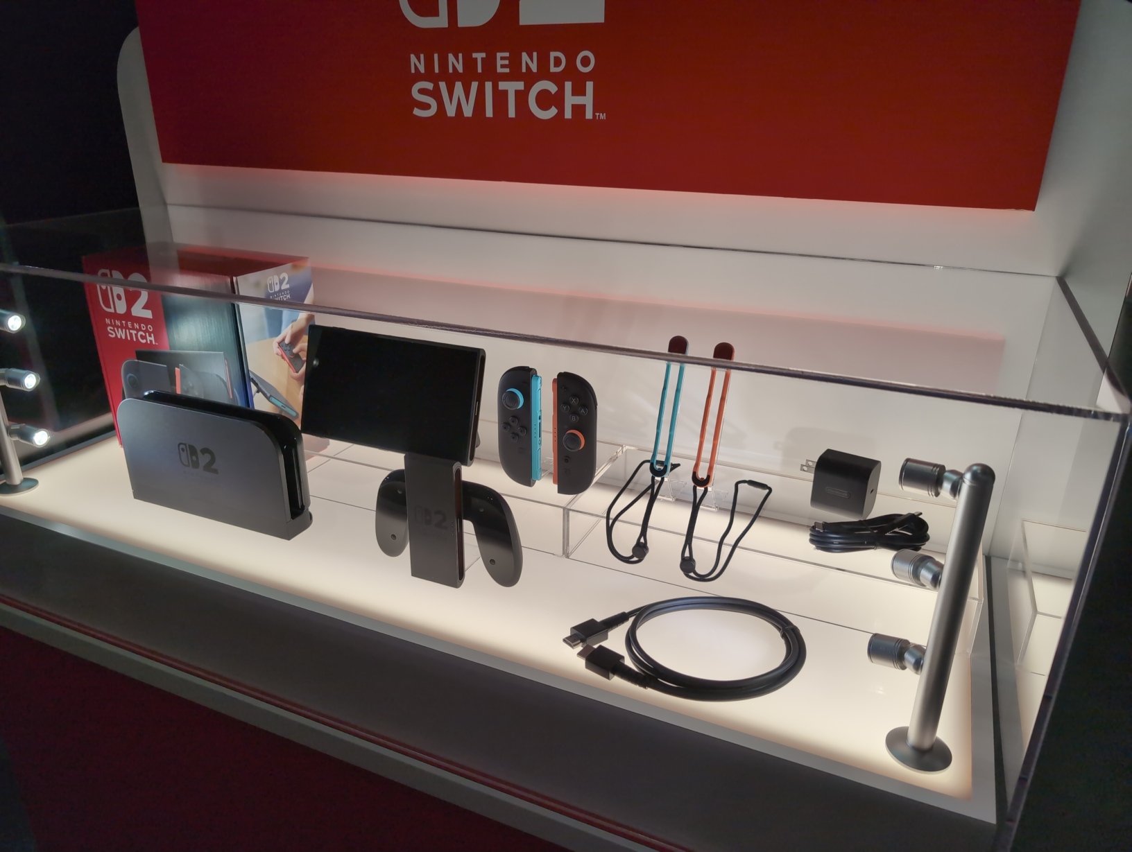 Photo of an illuminated display case showing a Nintendo Switch 2 box and all the hardware included inside: the console tablet, the left and right Joy‐Con 2 controllers and their wrist straps, the power brick and USB‐C power cable, the dock, the grip, and the HDMI cable.