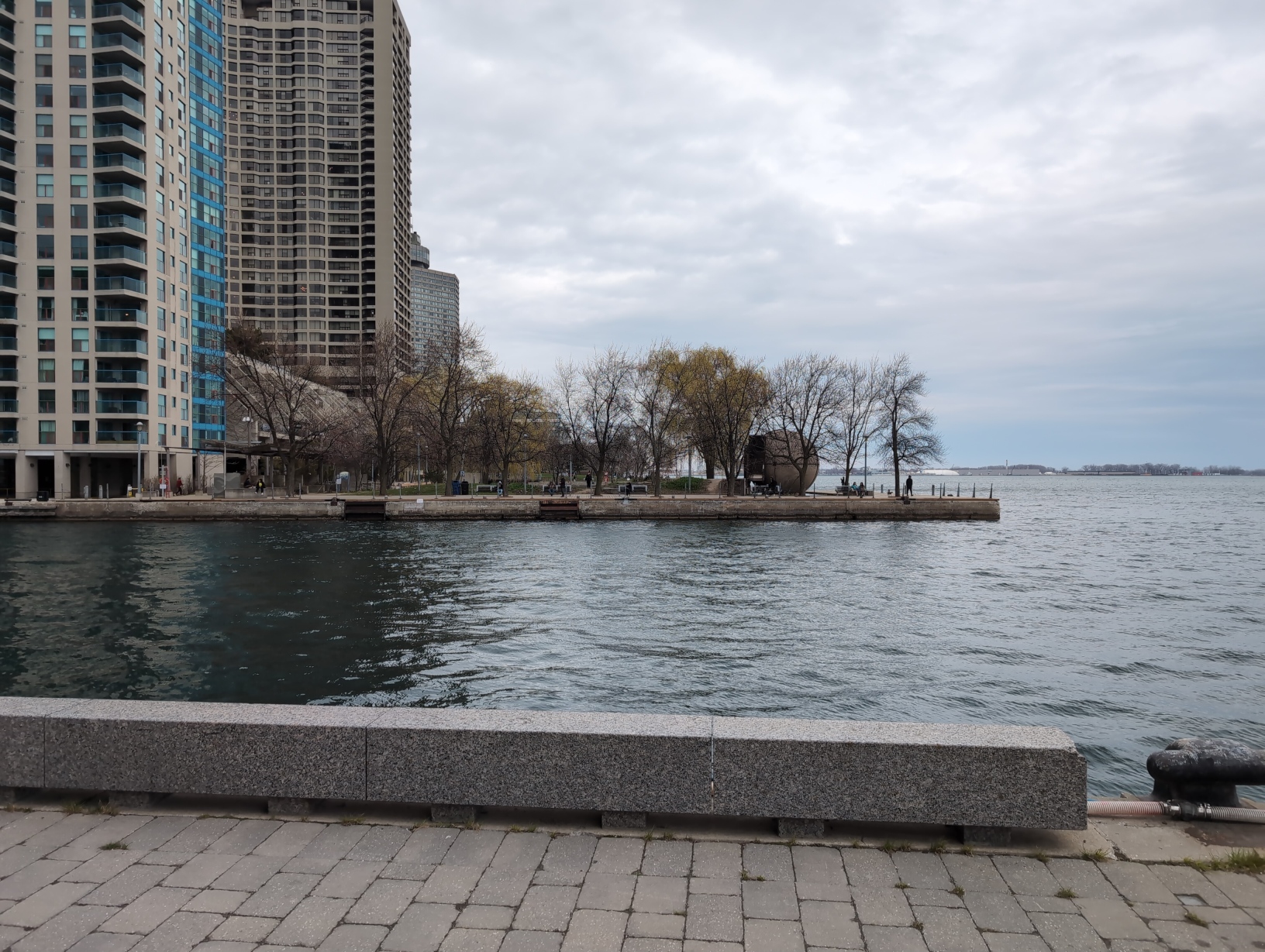 Photo facing Harbour Square Park West from the east, across the water. The left of the frame is filled by high‐rise condos; the centre by the park with grass and trees; and the right by the water of Lake Ontario, the overcast sky, and the distant trees and buildings at the horizon.