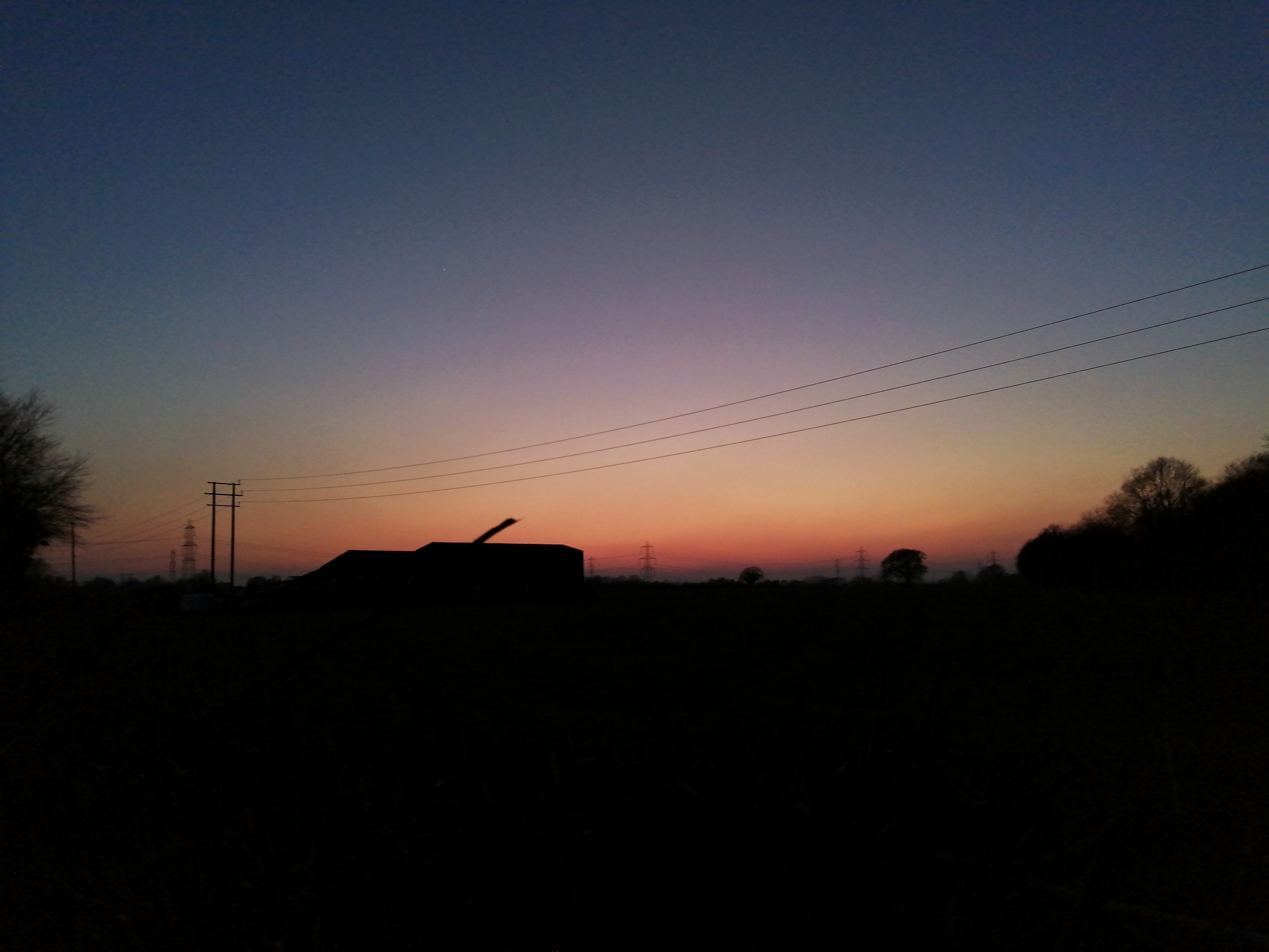 A sunset. The sky is blue at the top going onto bright orange. There are no details on the ground. We can only see the outline of trees, telephone lines, and a building