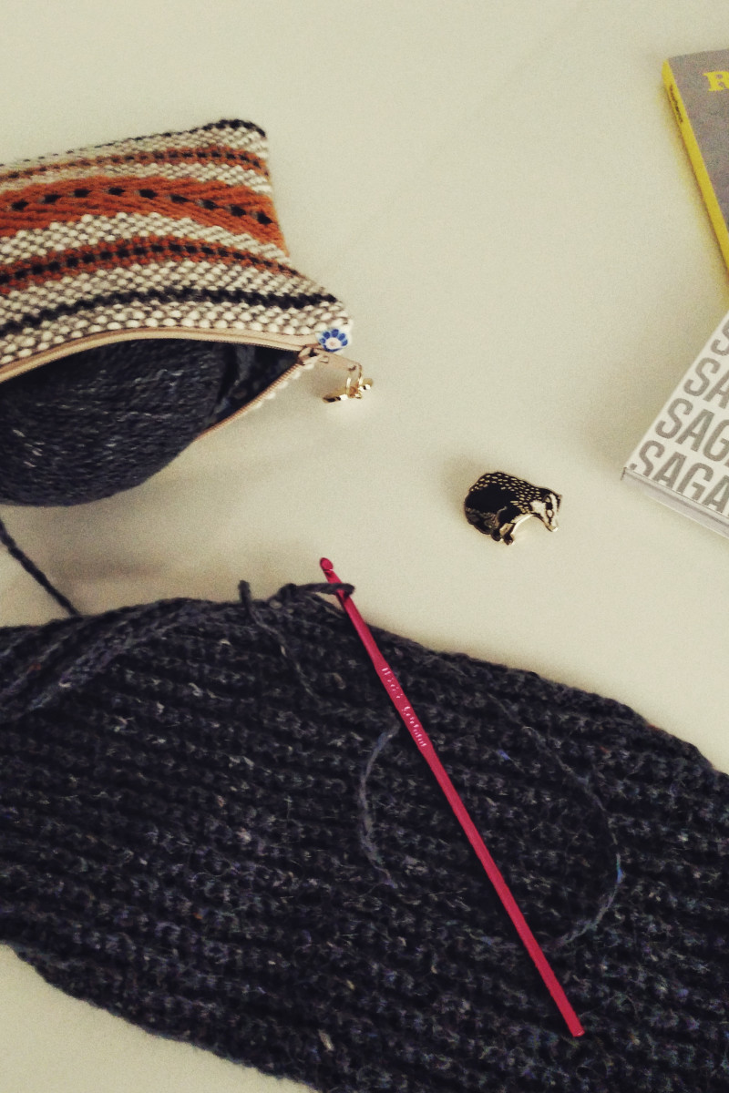 A vertical colour photograph of a crochet work in progress. The yarn used is grey. The work is cropped by the frame. It rests on a white table alongside a badger pin, a woven pouch, and the corner of a magazine.