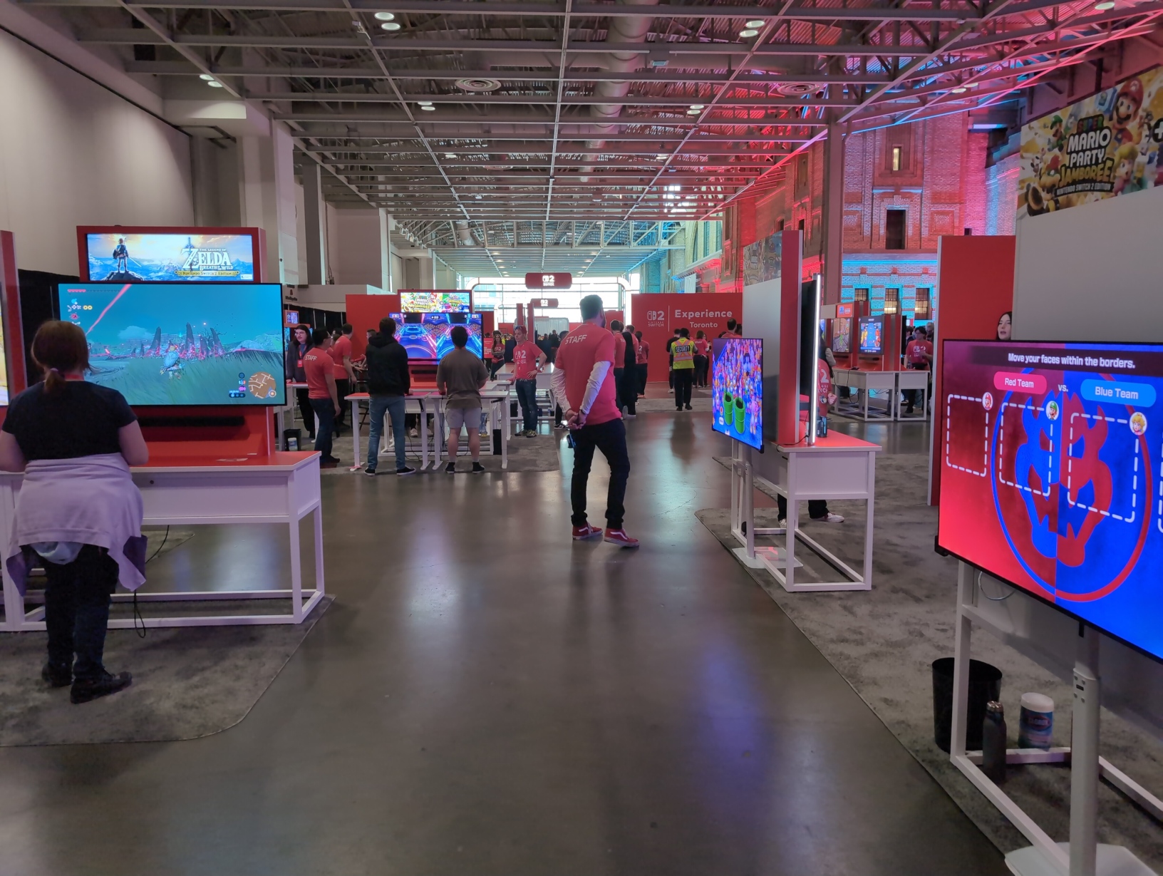 Photo looking across the free‐play area towards the entrance. Visible to the right are 2 setups for Super Mario Party Jamboree + Jamboree TV, where a cabinet has a TV set up on both sides, with one facing the public and the other facing towards a small booth with pure red walls. Beneath one of these cabinets sitting on the carpet are a small waste bin, a metal thermos, and a container of Clorox wipes. To the left is a setup for The Legend of Zelda: Breath of the Wild - Nintendo Switch 2 Edition.
