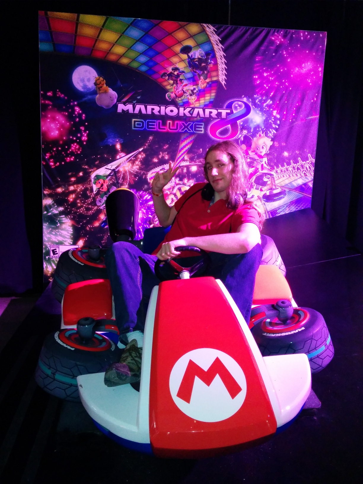 Photo of me in the photo op area for Mario Kart 8 Deluxe. The backdrop features the game’s key art and logo but without Mario in his kart, and instead a full‐scale model of Mario’s kart in anti‐gravity mode sits on a matte black floor in front under pink lighting. I’m sitting in the kart somewhat awkwardly as it’s too small for my tall frame, leaning to the side and smirking with one hand on the wheel and the other flashing a peace sign, and wearing a red polo, jeans, wireless headphones around my neck, and an attendee wristband.