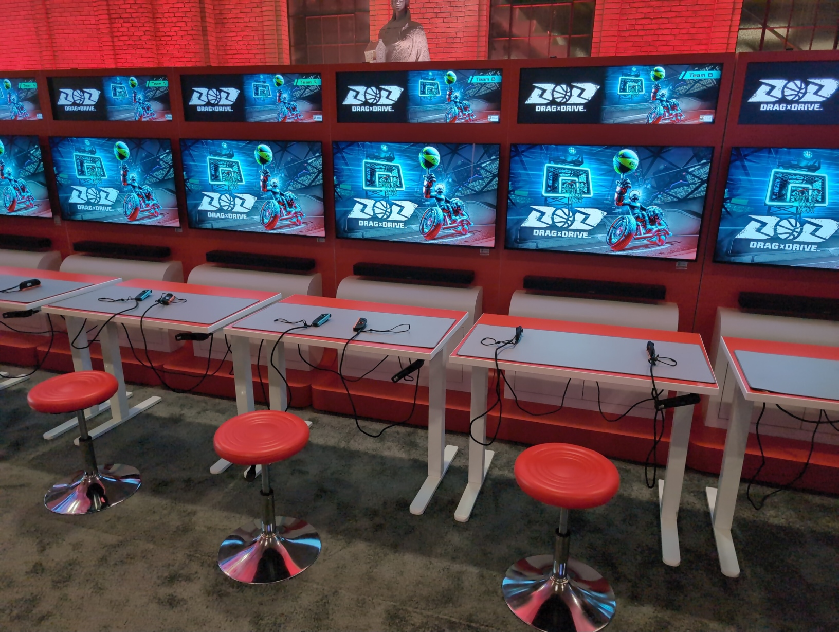 Photo of a row of 6 setups for Drag x Drive. Unlike other cabinets, these are more customized, solid units with no empty space below them, and the mounted TVs and soundbars are lower to the ground. The adjustable desks in front of these setups are all fully lowered, and have giant grey mouse pads and a set of Joy‐Con 2 controllers on them. In front of each desk is a metal stool with a round red plastic seat. Embedded in each cabinet above the TV is an illuminated sign with the game’s logo and key art, but also a label for “Team A” on the left 3 setups and “Team B” on the right 3.