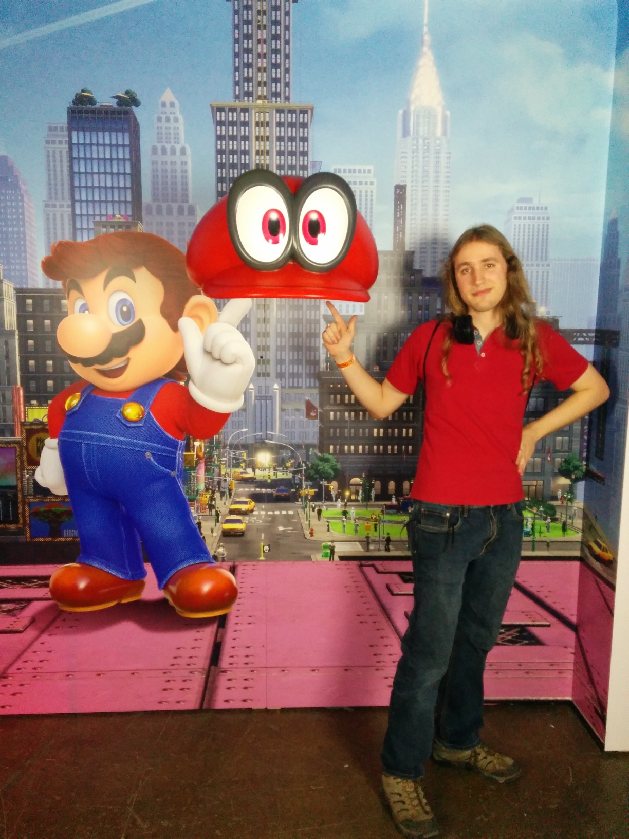 Photo of me in the photo op area for Super Mario Odyssey. The backdrop features Mario in the Metro Kingdom holding up a finger to his left but without Cappy twirling around it as usual, and instead a giant 3D model of Cappy is suspended at that position. I’m standing to the other side of Cappy mimicking Mario’s stance as I’m a head too tall to fit underneath it.