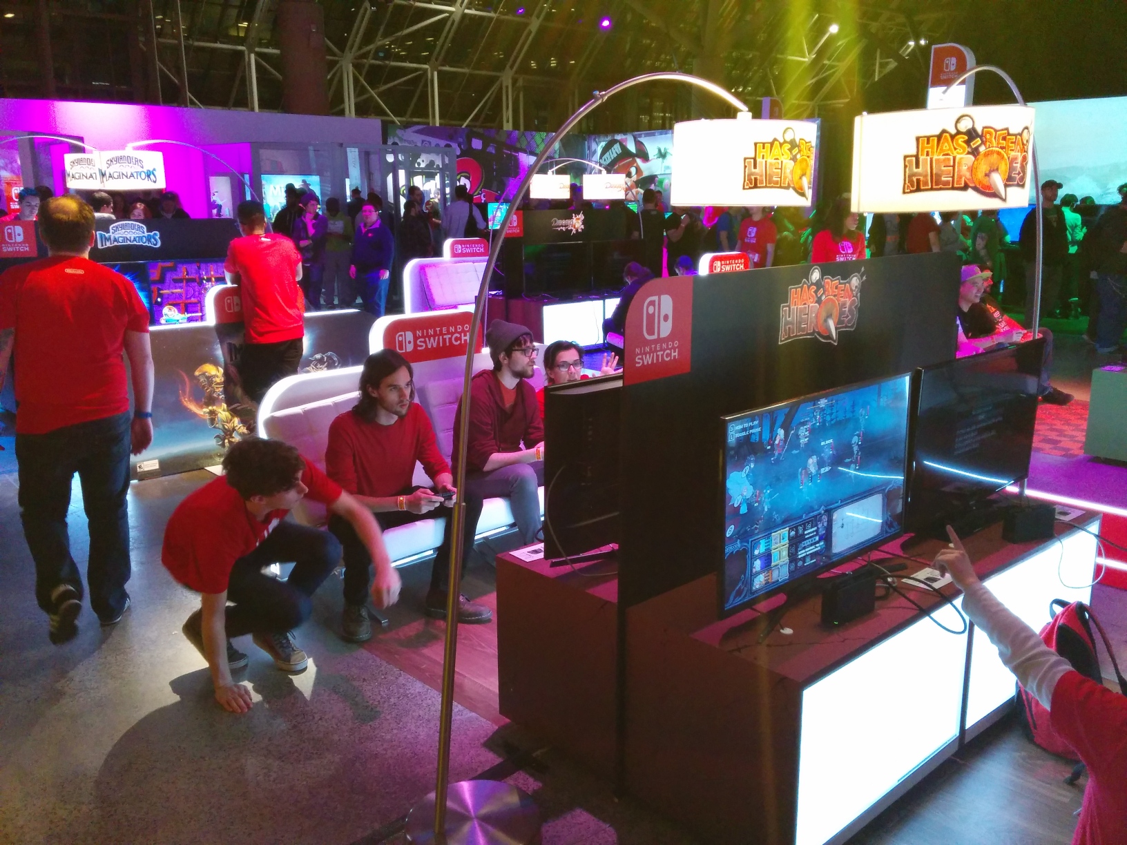 Photo facing various populated third‐party game setups in the central area. In front of each TV is some cushioned, reclined seating. Some staff members are sitting on the floor next to seats occupied by attendees.