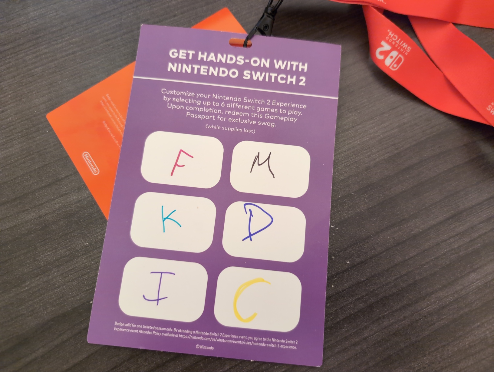 Photo showing the reverse of the Gameplay Passport. On a purple background, there’s a header, a short blurb, 6 rounded white squares taking up most of the space, and 2 lines of fine print. The squares are all filled in with letters drawn with coloured permanent markers, with most of them being fairly messy: a red “F”, a black “M”, a cyan “K”, a blue “D”, a purple “I”, and a yellow “C”. The header reads “GET HANDS‐ON WITH NINTENDO SWITCH 2”, and the blurb reads: “Customize your Nintendo Switch 2 Experience by selecting up to 6 different games to play. Upon completion, redeem this Gameplay Passport for exclusive swag. (while supplies last)”