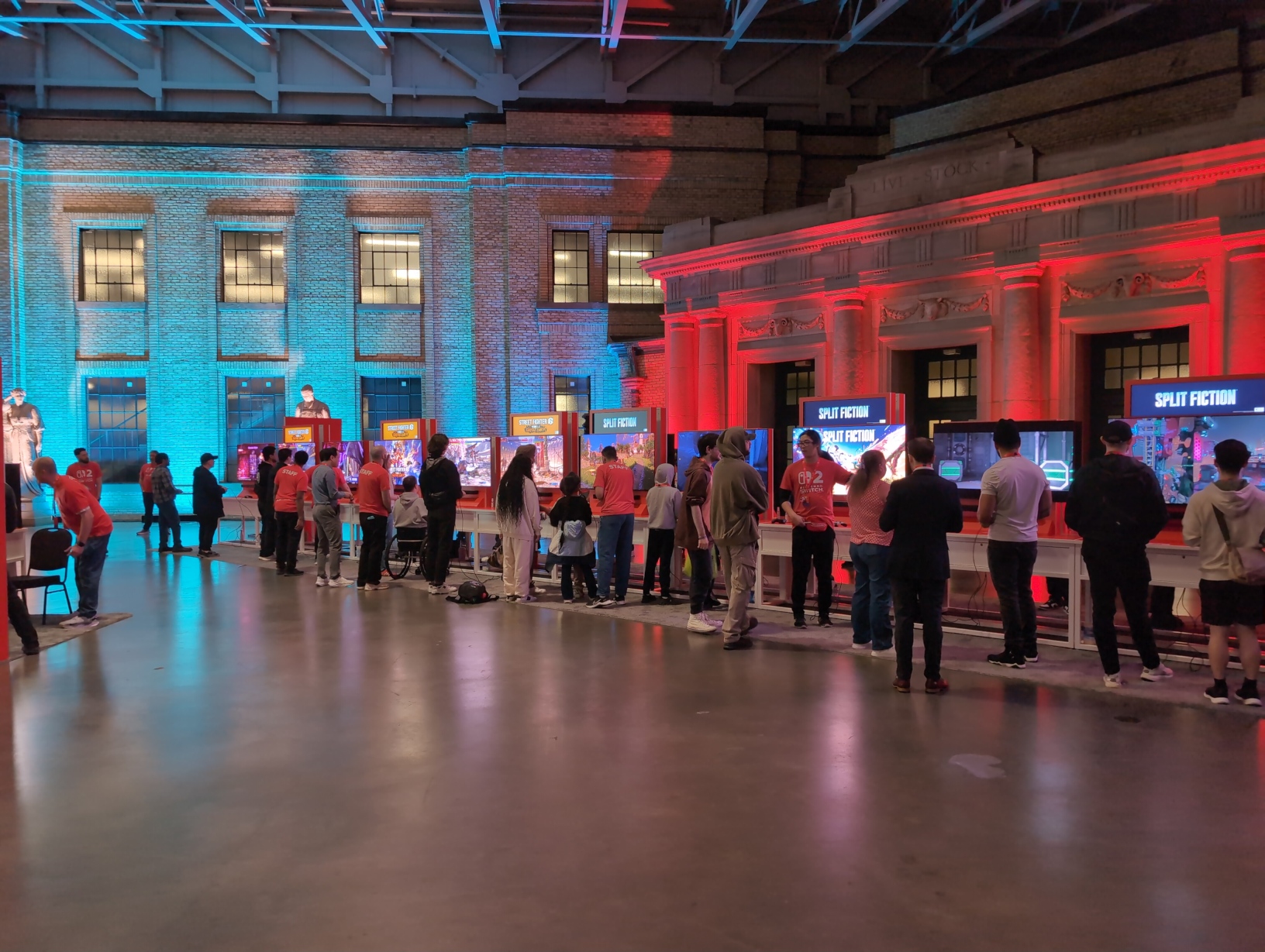 Photo of a very long row of populated setups at the edge of the free‐play area, with 6 for Street Fighter 6 and 6 more for Split Fiction. Distantly visible a staff member in the open area to the left of all these setups, standing in a wide stance facing towards the rest of the free‐play area with their hands in their pockets, clearly just keeping watch.