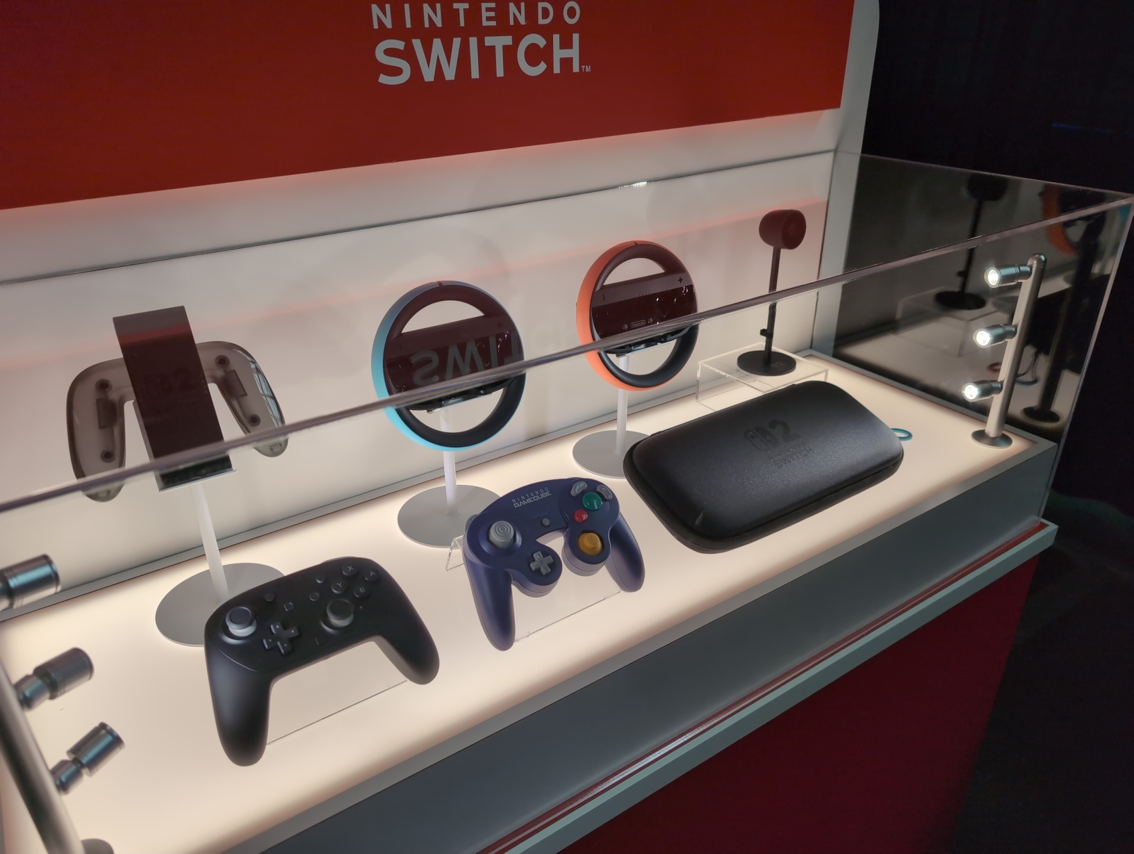 Photo of an illuminated display case showing official Nintendo Switch 2 accessories on stands: the charging grip, the blue and red Joy‐Con 2 wheels, the camera, the Pro Controller 2, the new wireless GameCube controller, and the carrying case.