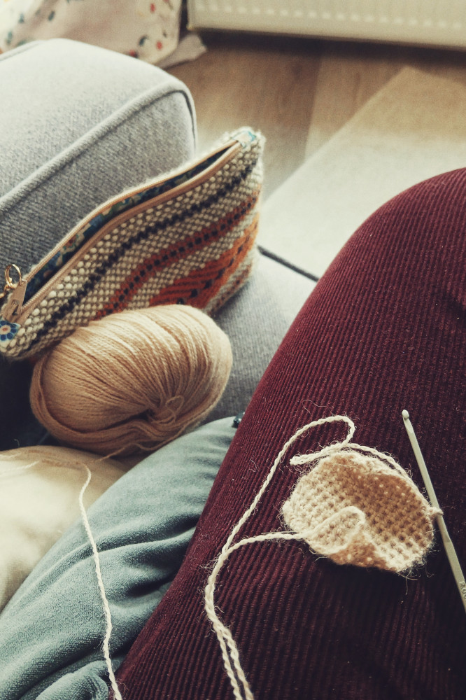 A vertical colour photograph of a small crochet work on my leg. I am wearing burgundy corduroy. The crochet work is a small rectangle made with creamish yarn on a small grey hook. I am on a sofa, the edge just visible. The yarn ball rests against the armrest as well as a woven small pouch.