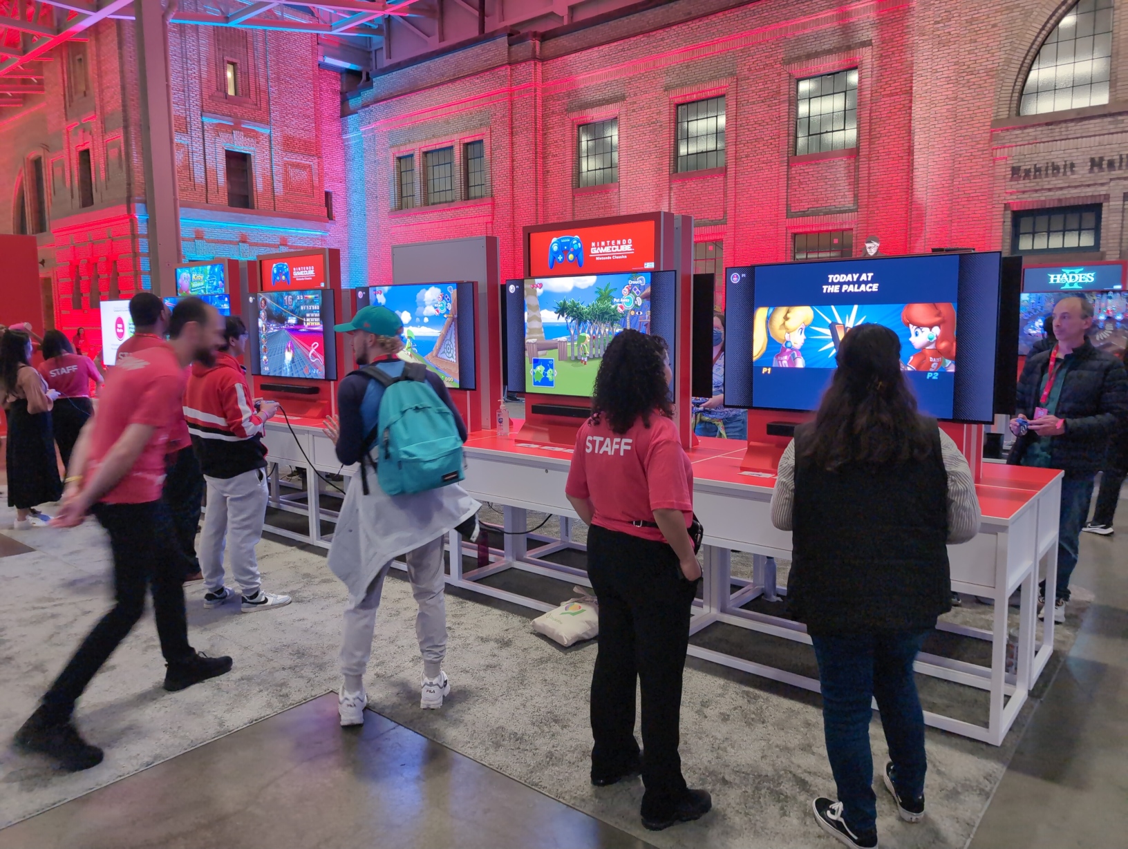Photo of a row of stations in the free‐play area, consistently mostly of Nintendo GameCube: Nintendo Classics setups. Of those, 1 is running F‐Zero GX, 2 are running The Legend of Zelda: The Wind Waker, and 1 is running Super Mario Strikers. The brick walls in the background are illuminated from below by red and blue spotlights.