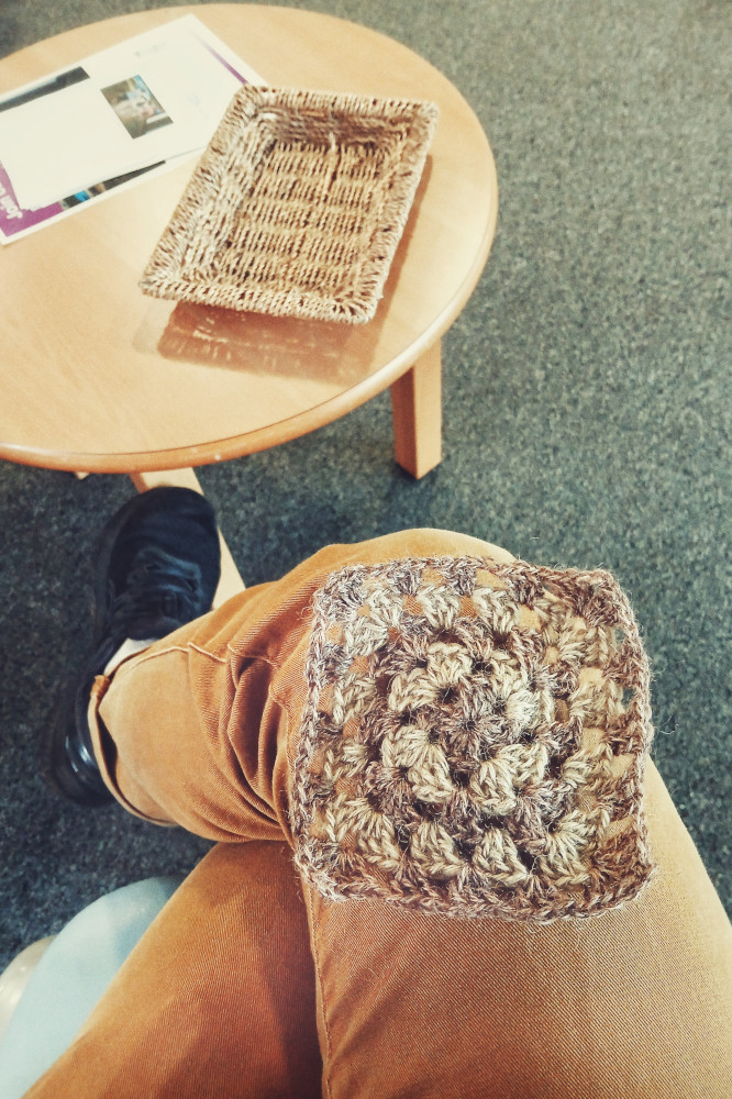 A vertical colour photograph of a brown and green granny square resting on much crossed legs (I'm wearing mustard coloured jeans). Around my legs are a grey waiting room carpet, a round coffee table with some leaflets and a woven empty rectangular tray.