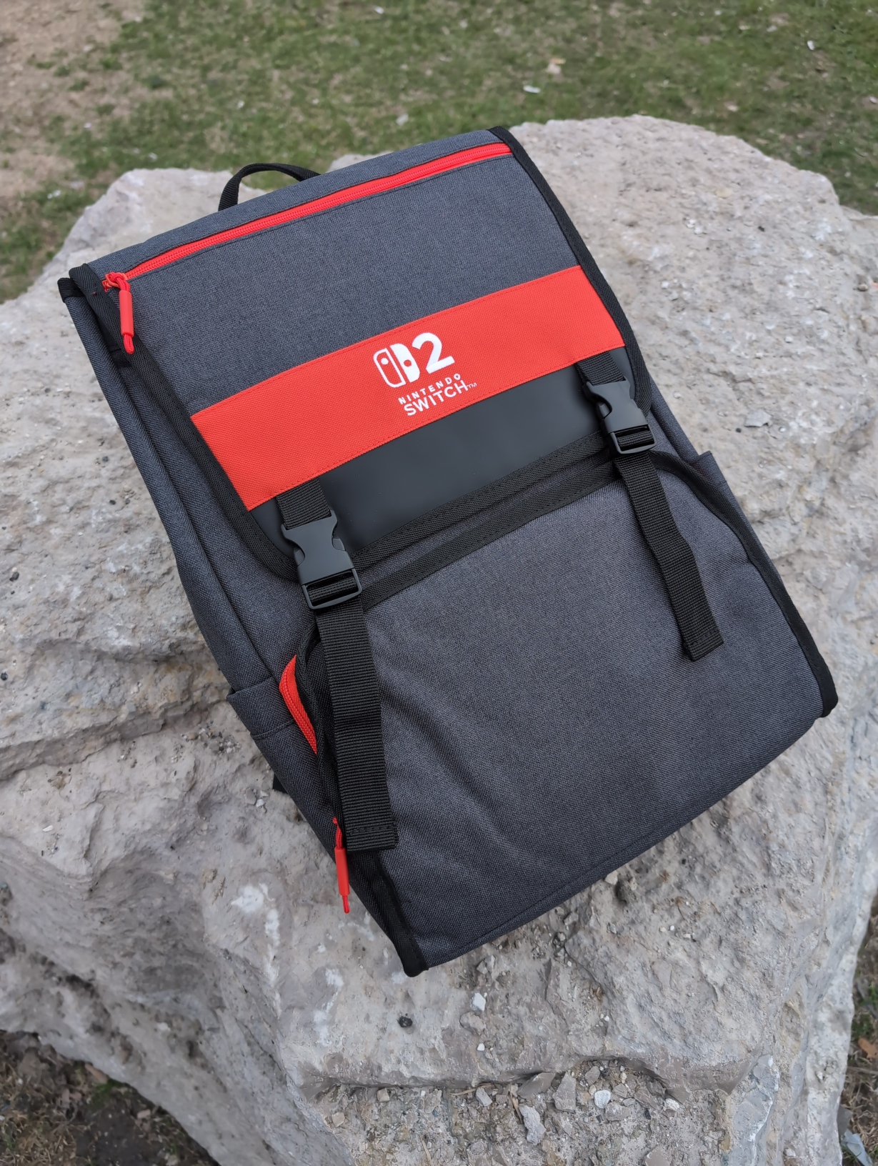 Picture of a bag resting on a large rock outdoors. It’s fairly squared‐off in shape and is dark grey with red accents. A big red stripe with the Nintendo Switch 2 logo stitched in the middle goes across a flap covering half of the bag’s front which is held down by 2 plastic buckles. There’s a zipper pocket occupying the bottom half of the bag’s front, as well as a red zipper at the top for a pocket in the flap itself. Open pockets at the bottom of each side are also visible, well‐sized for holding a water bottle.