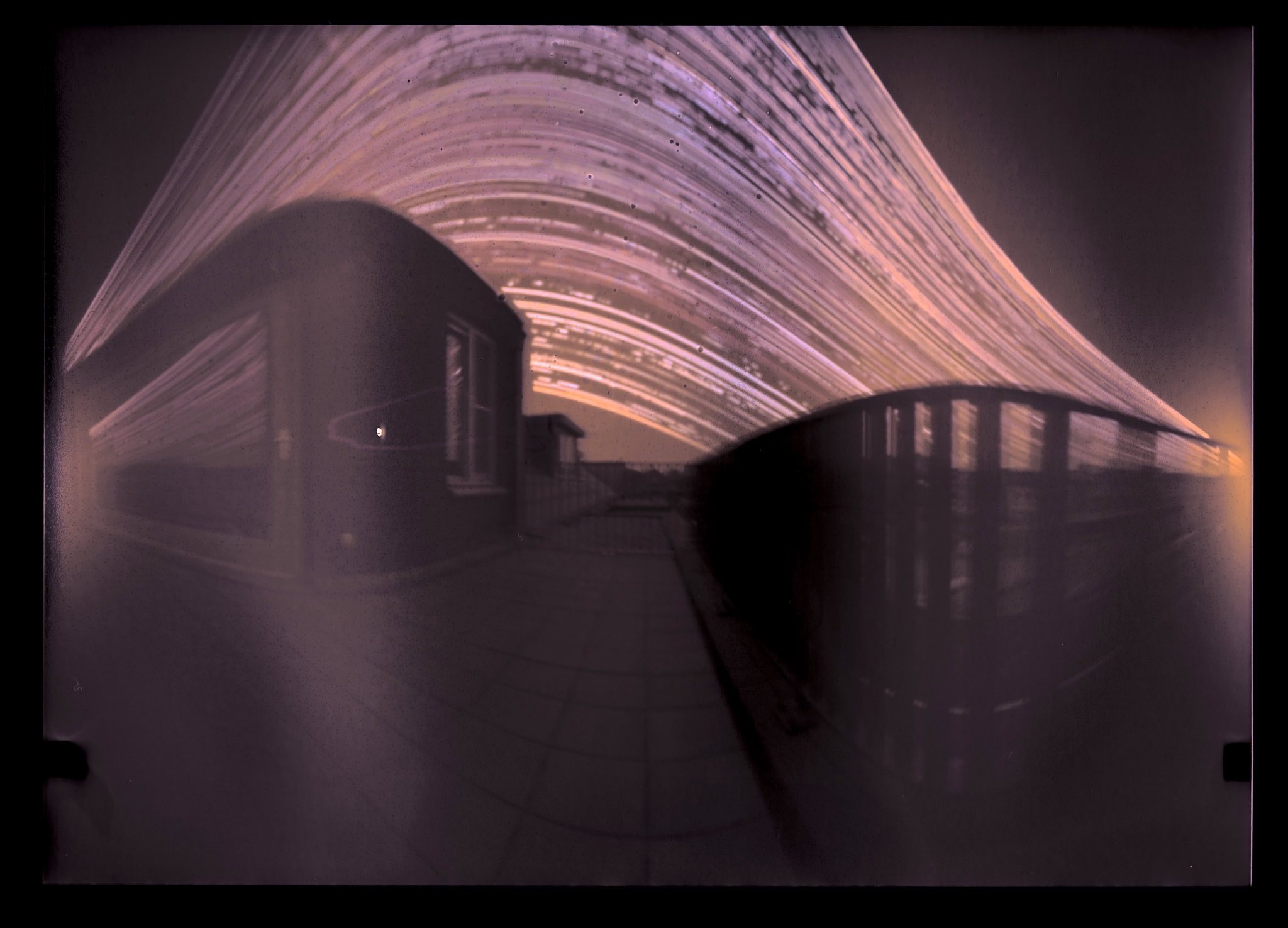 This image shows a long-exposure photo capturing sub trails in the sky above a building rooftop and a fence.