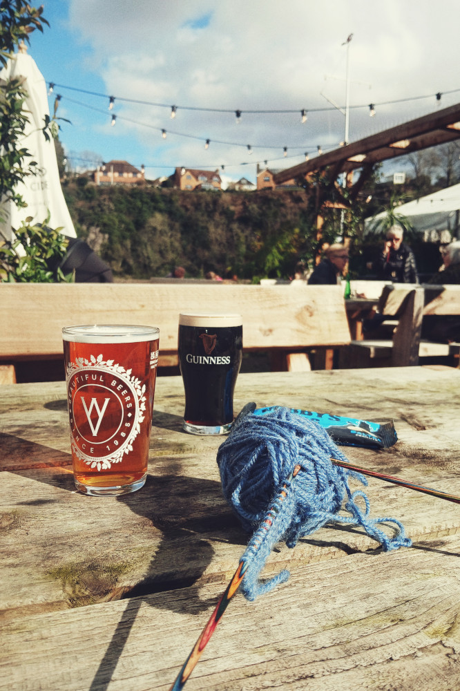 A vertical colour photograph of an outdoor pub table with two half pints on it (a Guinness and a light ale), a packet of nuts, and a blue ball of yarn with two colourful wooden knitting needles stuck in it.