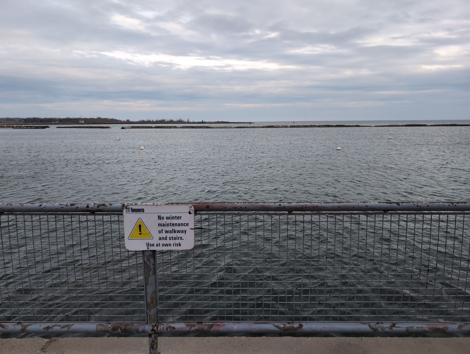 Photo of Lake Ontario on an overcast day, from behind a metal fence with a Government of Toronto warning sign which reads: “No winter maintenance of walkway and stairs. Use at your own risk.” The vast expanse of blue‐grey water is broken up only by some barriers, buoys, and the Toronto Islands occupying the left half of the horizon.