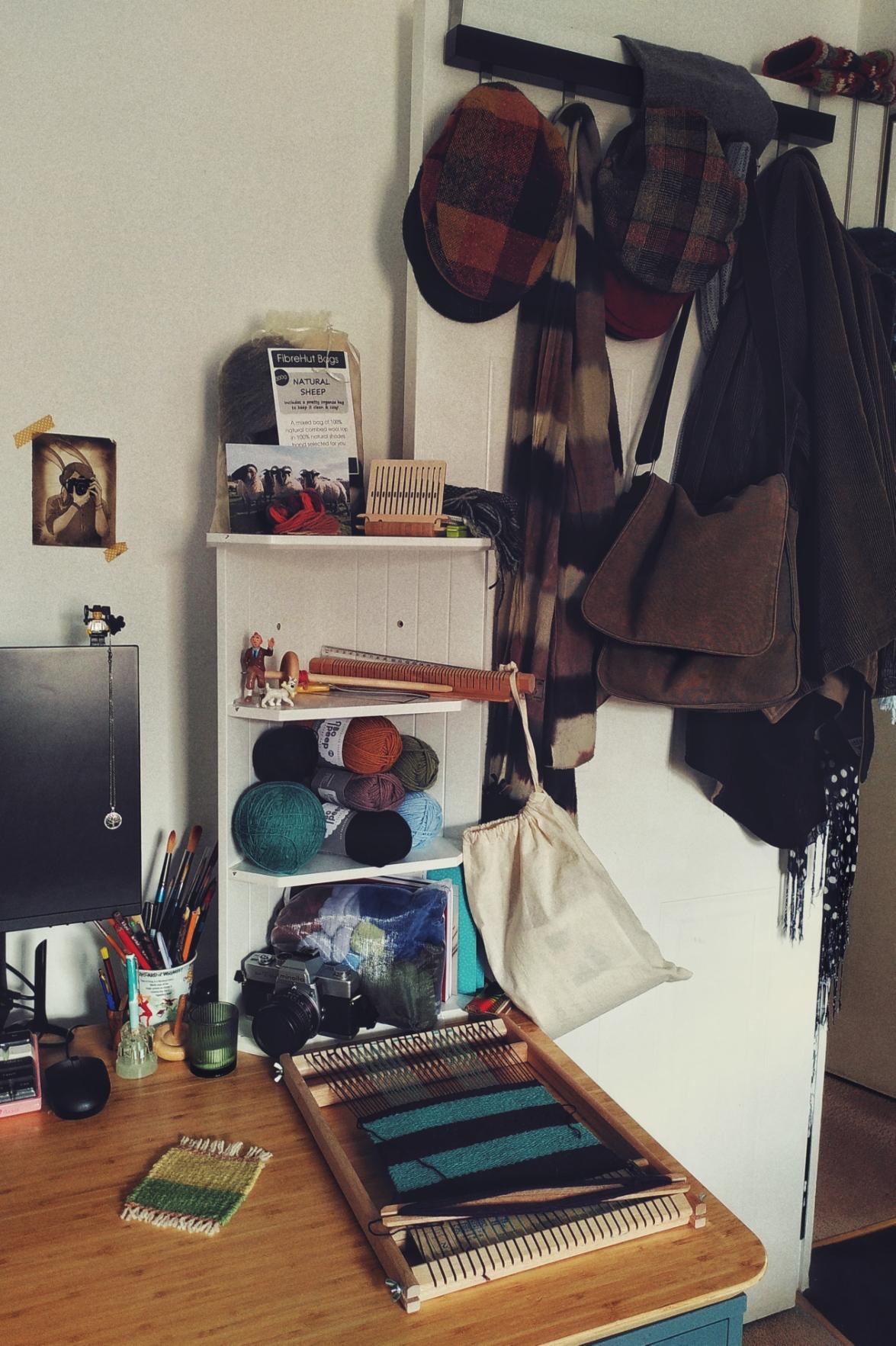 A vertical colour photograph of a desk corner with a small storage that holds fibre, wool, and associated weaving and spinning tools. In front of it is a loom with a turquoise and purple weave in progress. Next to the desk is a white door that holds an array of wooden flat caps, jackets, scarves, and a canvas bag.