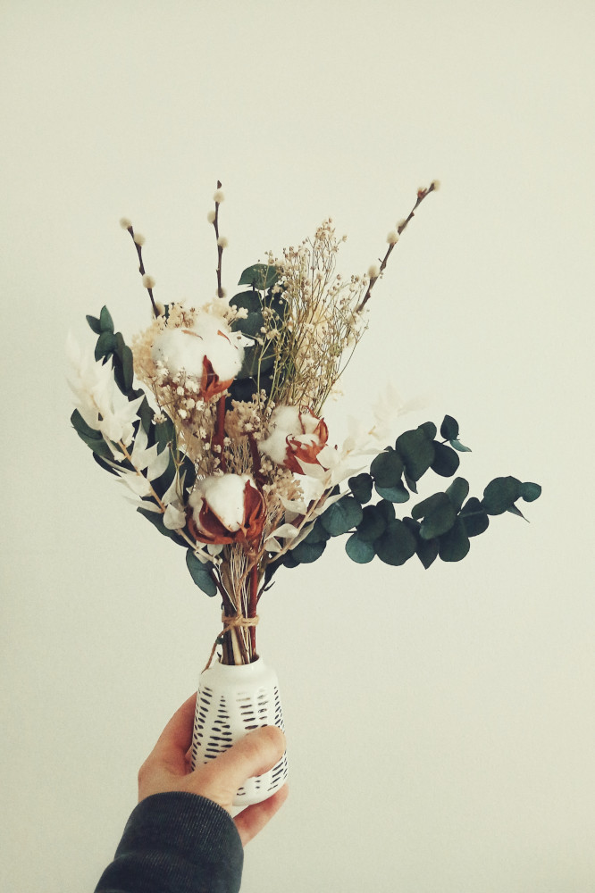 A vertical colour photograph of my white hand holding a small white vase with streaks of blue. In it are dried flowers including eucalyptus and cotton.
