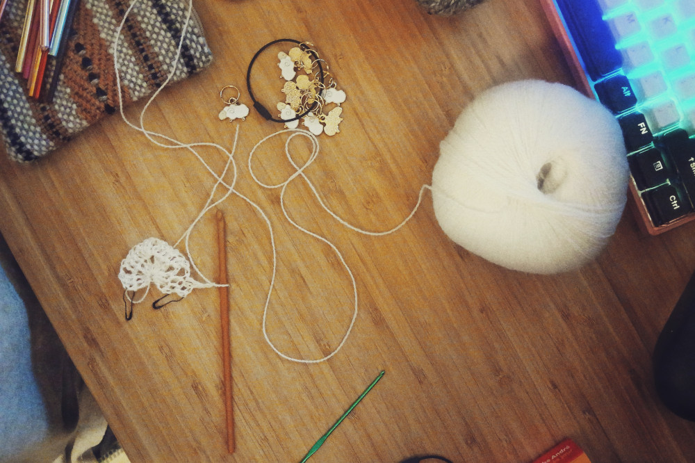 A horizontal colour photograph of a bamboo desk with the start of a crochet work on it. The hook is attached to it. A trail of yarn leads back to the ball. It's white. Around are a keyboard, sheep shaped stich markers, and the corner of a woven pouch.