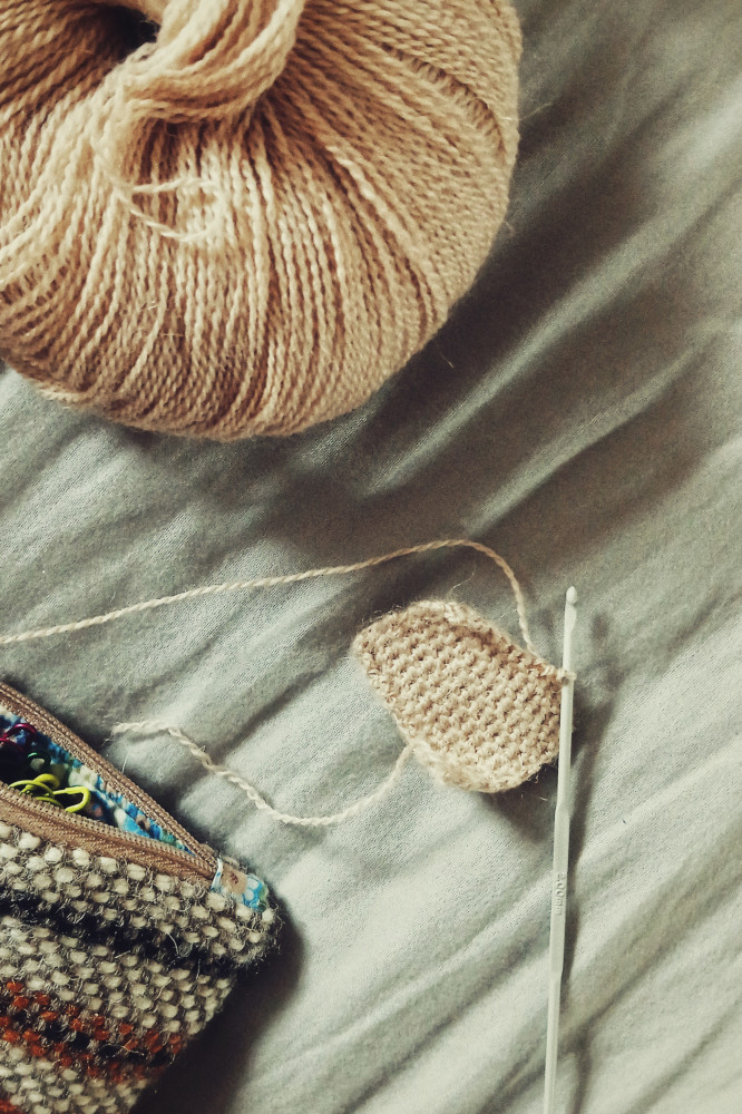 A vertical colour photograph of a very small crochet work. It's so far a rectangle on a 2mm grey hook. The yarn is of a creamish colour. The ball is at the top left corner of the image. At the bottom left is a corner of a woven pouch with grey, black, and orange colours on it.