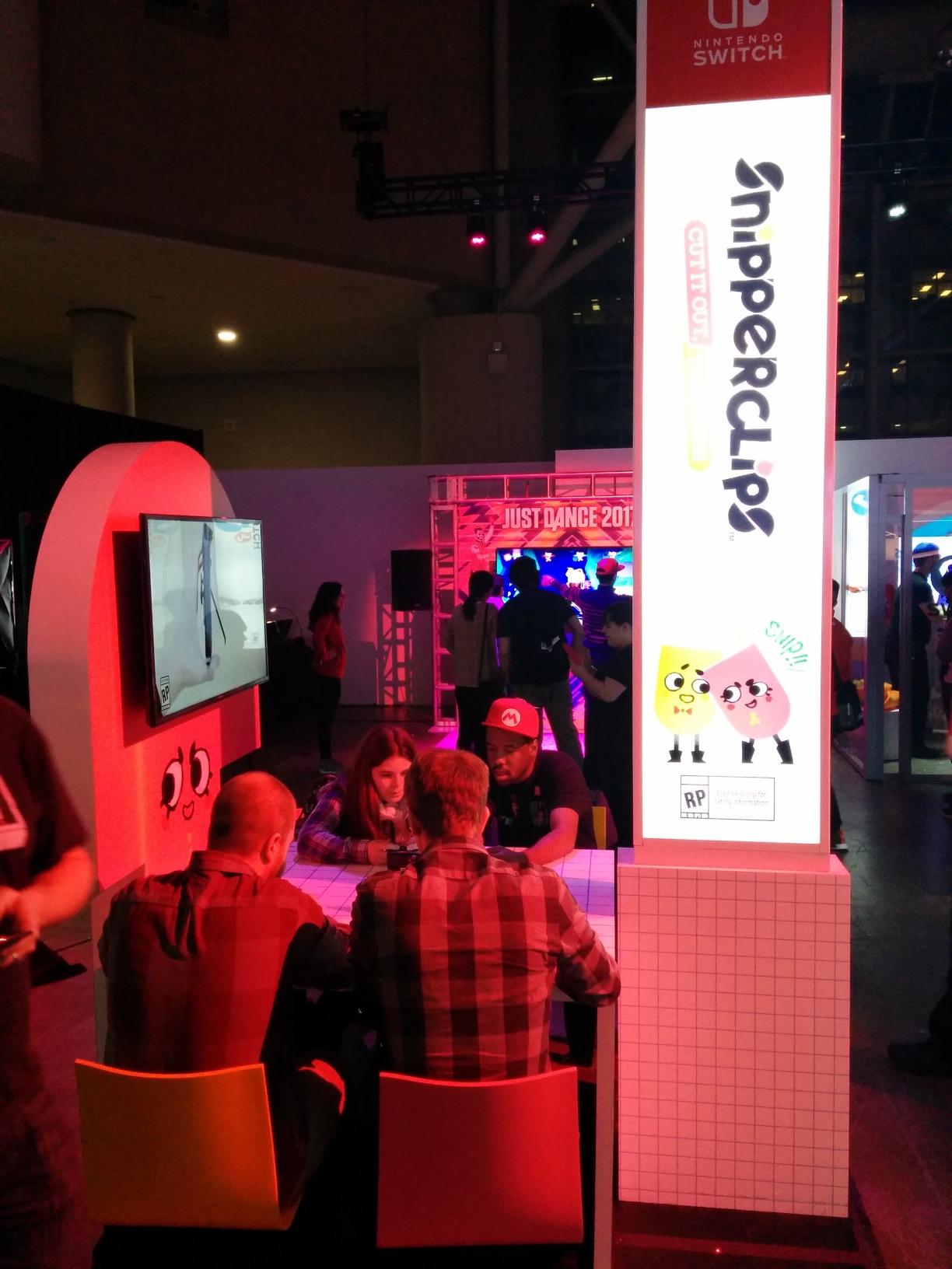 Photo of the Snipperclips area. Between a tall illuminated sign for Snipperclips and a TV mounted on a statue resembling Snip is a table with a “grid paper” design, holding 2 “tabletop mode” Switch setups. It seats 2 attendees on each side, with each having 1 yellow and 1 pink chair. Behind this, a setup for Just Dance 2017 is visible, with the TV mounted on a backdrop of key art surrounded by faux steel stage scaffolding with mounted speakers.