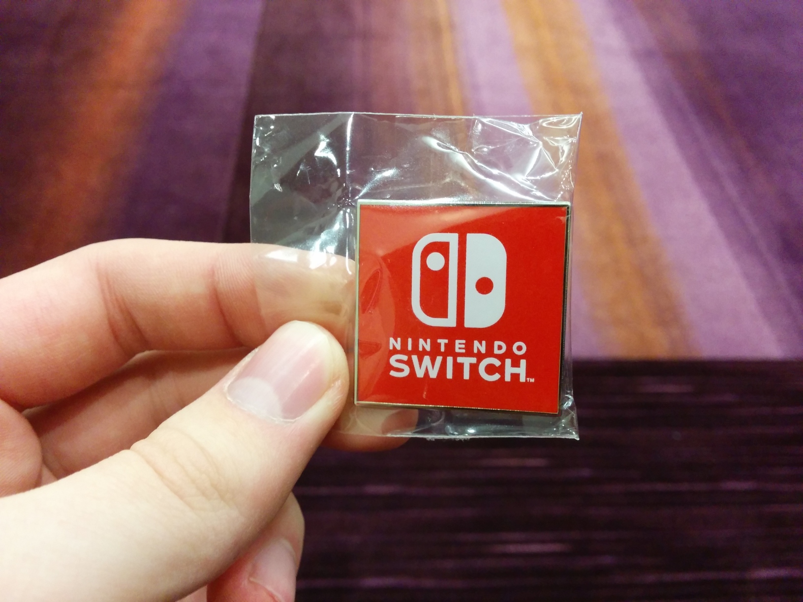 Photo of my hand holding a square enamel pin in plastic wrap. It’s simply filled with a white Nintendo Switch logo on a pure red background.