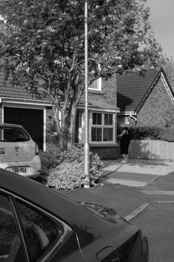 A vertical black and white photograph of a woman tinkering in her front garden made of gravel. She is in the background in a residential street. In the foreground is the back of a dark car creating distance between the viewer and the woman