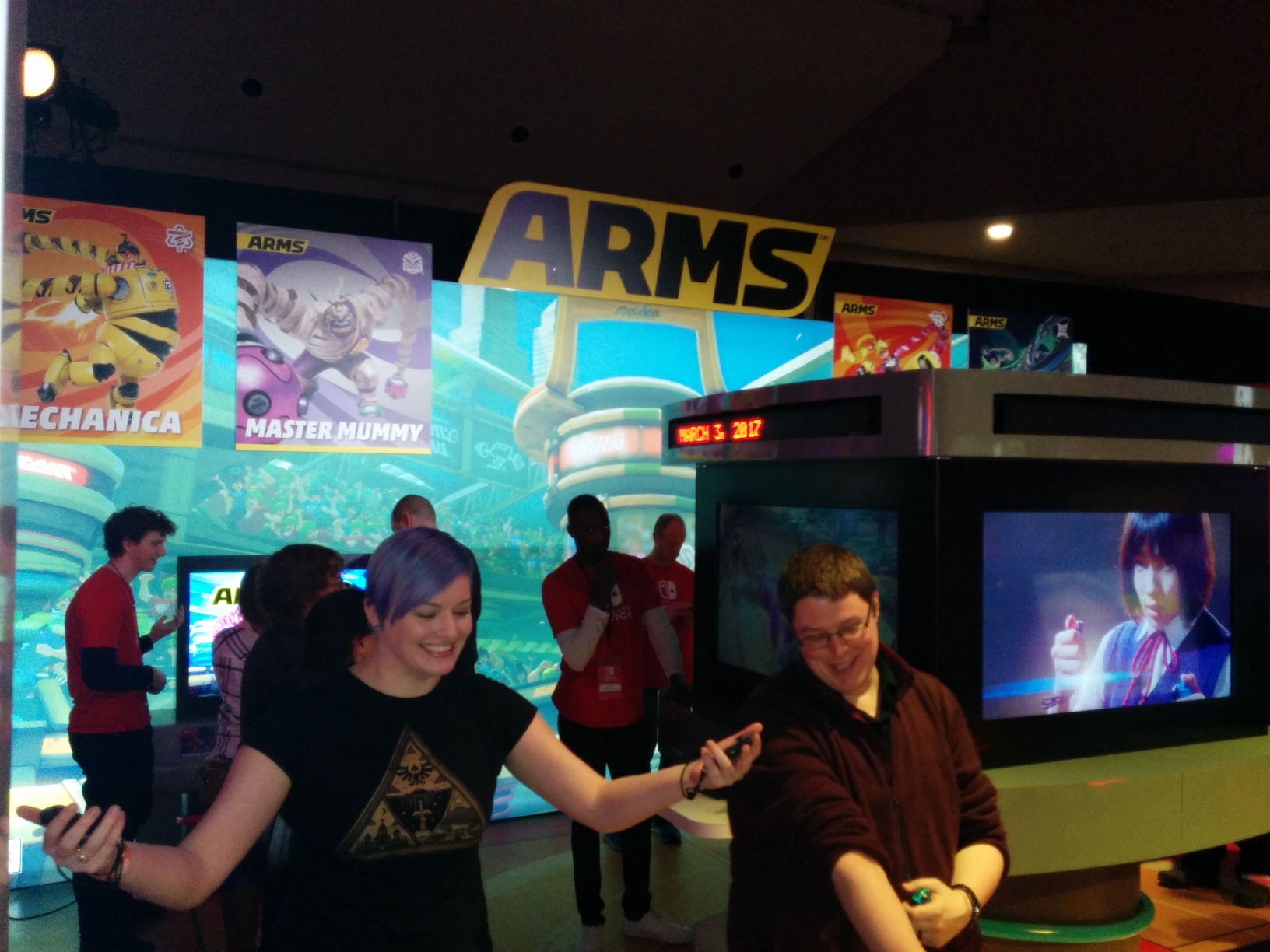 Photo of the ARMS area. A central console has a TV covering each of its 4 faces, with 1 playing a promotional video and the others hosting game setups. Scrolling LED displays go around above these TVs, and currently visible on one is the Switch’s launch date: “MARCH 3, 2017”. Behind it is a backdrop of Spring Stadium with more demo setups in front, and hanging above are a giant ARMS logo and 4 banners with key art and names of some of the game’s characters: Mechanica, Master Mummy, Ribbon Girl, and Ninjara.