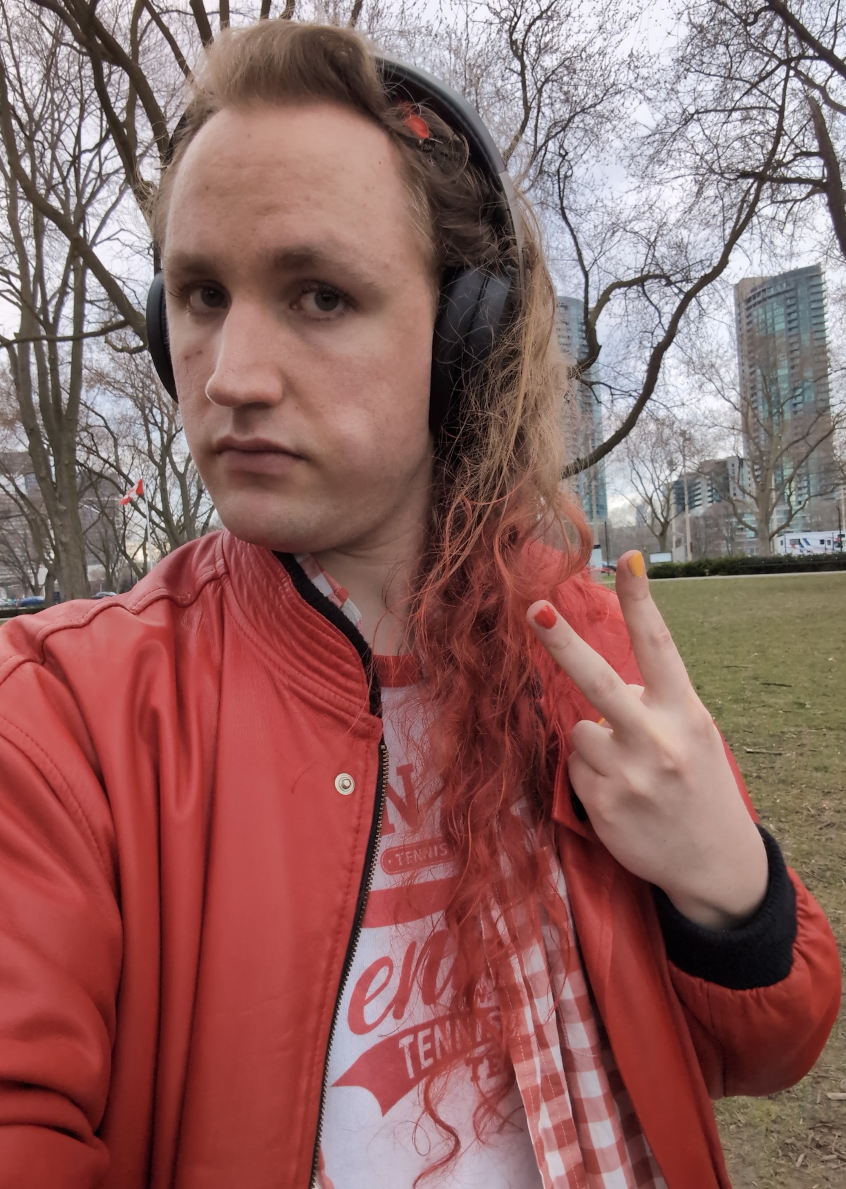 Selfie taken in Coronation Park. I’m wearing over‐ear headphones on my head of long wavy blonde hair, which is dyed red below my shoulders. My expression is neutral, with my face tilted somewhat away from the camera to let my hair down over my far shoulder. I’m flashing a peace sign at the camera, and the index and middle fingers are painted red and yellow respectively. I’m wearing a red leather jacket, an unbuttoned red and white plaid button‐up, and a white t‐shirt with a red graphic that just seems to be the word “tennis” in a bunch of different fonts.