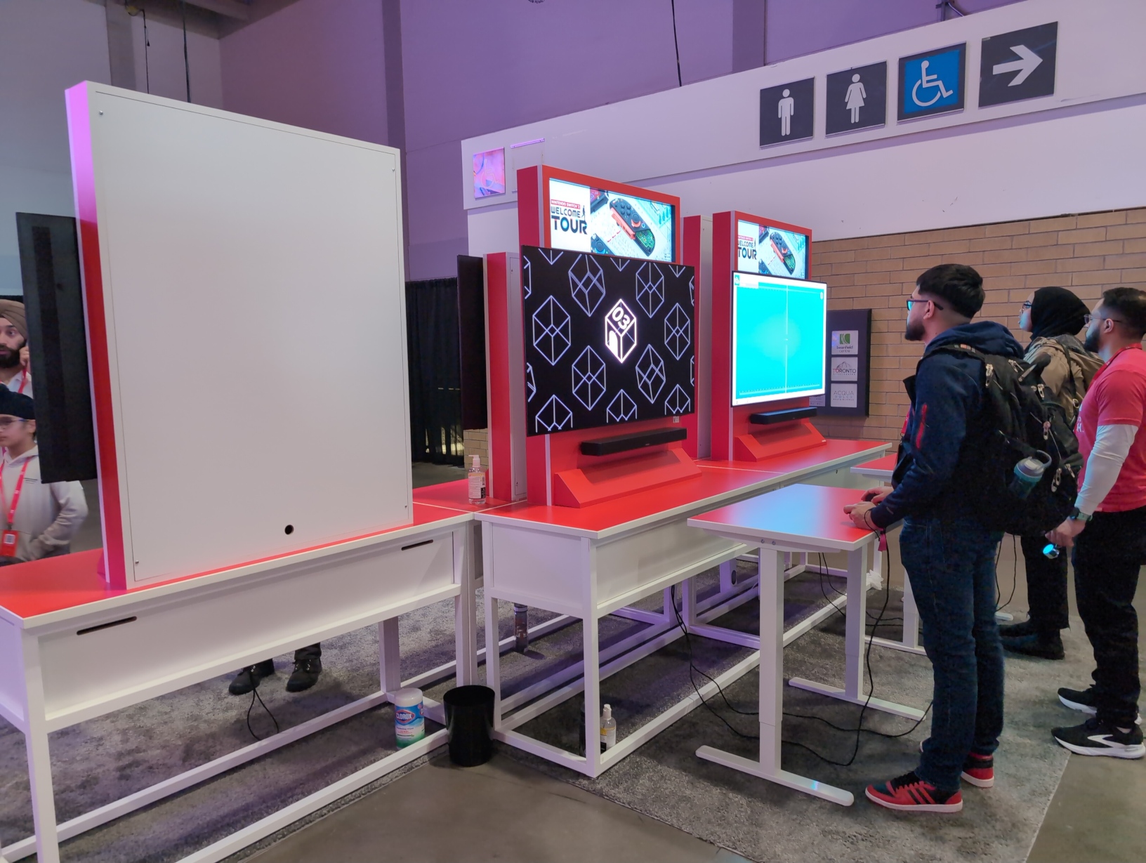 Photo of 2 setups for Nintendo Switch 2 Welcome Tour. Both have players at them. They are up against 3 more setups facing the other direction, but there’s no third setup for this game, so the empty backside of one cabinet is visible next to these 2 setups. Beneath these cabinets sitting on the carpet are a small waste bin, a metal thermos, a bottle of hand sanitizer, and a container of Clorox wipes.