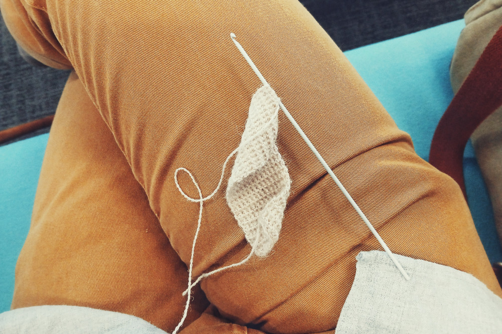 A horizontal colour photograph of my legs in mustard jeans with a rectangle strip of crochet work in progress. The work is made with a creamish coloured yarn on a small grey hook. I am seated on a sky blue sofa.