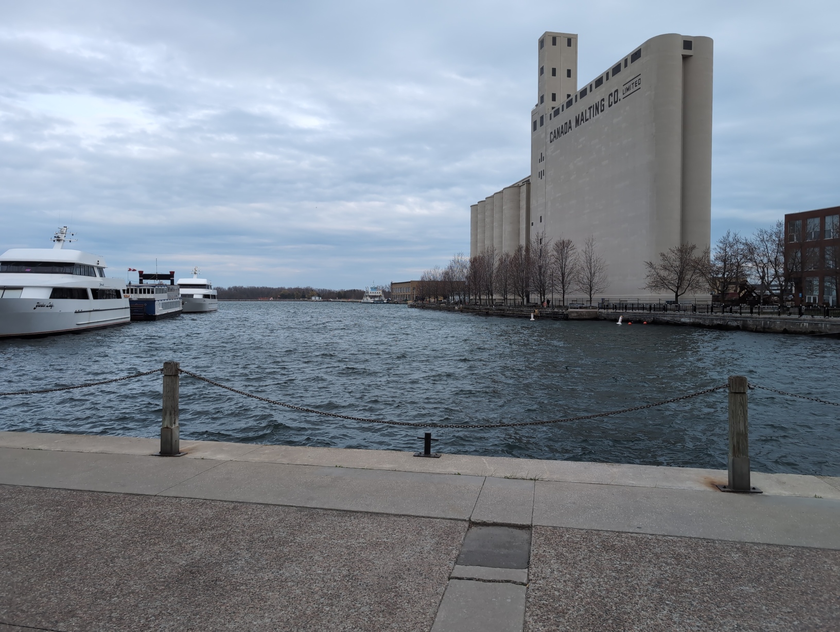Photo facing Lake Ontario from the harbourfront. On the left side of the frame are 3 docked tour boats, 2 with 2 levels. The right half has a protruding landmass with a tall, prominent, concrete building with several silos labelled “CANADA MALTING CO. LIMITED”, which looks very imposing with its sheer faces of pure concrete interrupted only by the aforementioned giant label and sparse blacked‐out windows. Visible between these across the water are the Toronto Islands.