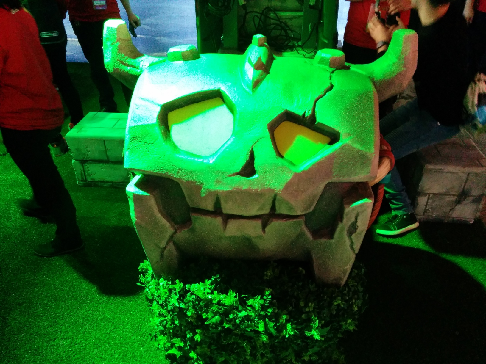 Photo of a statue of an outpost chest in the exhibition area for The Legend of Zelda: Breath of the Wild at the Nintendo Switch Preview Tour. It resembles a skull and sits on a fake hedge, itself on the floor covered on faux turf grass. Behind it, attendees and staff surround TV setups with faux brick stools. The scene is dominated by green lighting.