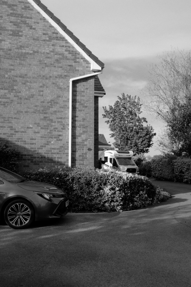 A vertical black and white photograph of houses drives with a car and a motorhome parked. The car is in the foreground, dark, and with only the front wheel and bonnet of the car visible. It is in front of a brick house. The motorhome is in the background next to the house. There a
is a leafy bush between it and the car. On the left of the image are more bushes and trees. Light streams through in patches.
