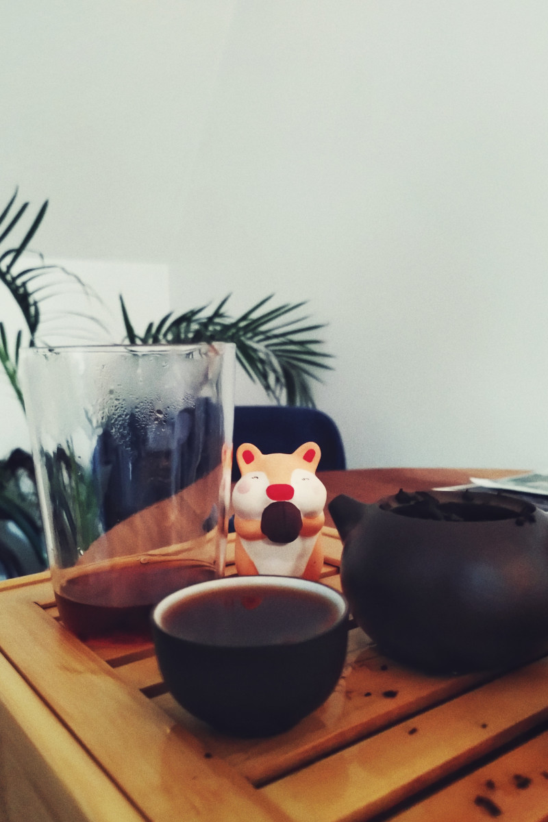 A vertical colour photograph of a gong fu tea set on a bamboo tea tray. Amidst the clay and glass tool is a cute hamster tea pet.
