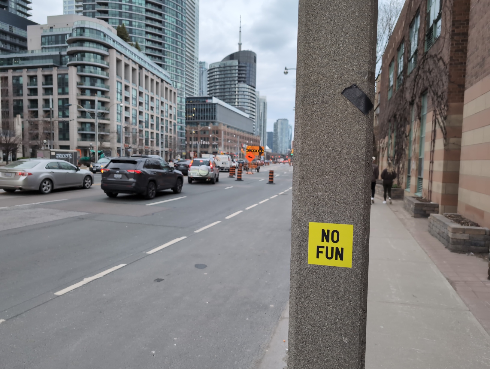 Photo from the sidewalk looking down a busy street with various construction signs and pylons obstructing most lanes of traffic. Everything is somewhat out of focus except for the concrete post occupying the centre‐right of the frame, which is adorned with a yellow sticker with black text that simply reads “NO FUN”. The view of the grey, cloudy sky above the traffic is mostly obstructed by glass high‐rise buildings.