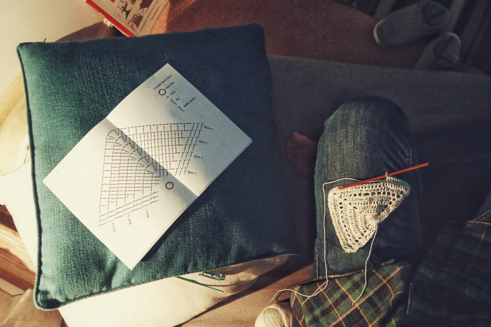 A horizontal colour photograph of a triangular small white crochet work on my knee. I'm wearing blue jeans. A crochet hook is attached to the work. Next to me.om the sofa are two pillows. The top one is teal and holds an open paper with a crochet pattern diagram on it. The ball of yarn is nestled by my side.