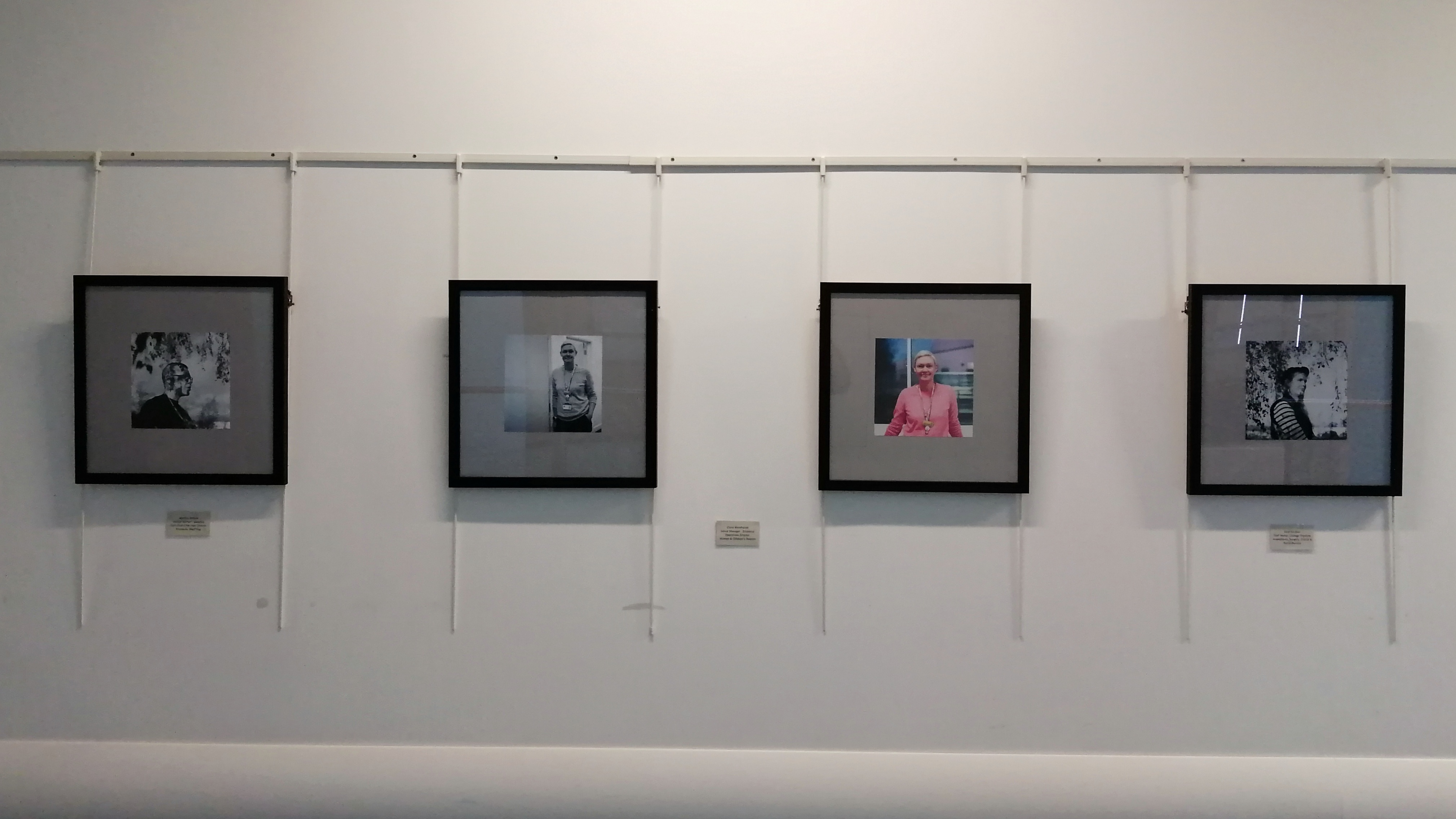 Four picture frames with portraits on them. Three portraits are in black and white. One is in colour. They are hung on a white wall.