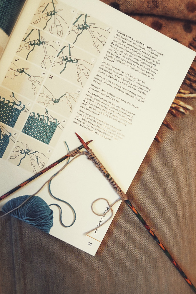A vertical colour photograph of a book about learning to knit. Two colourful wooden knitting needles rest on it with the very beginning of a knitting something on them.