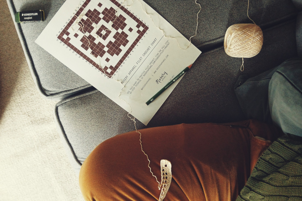 A horizontal colour photograph of a frogged filet crochet. The thread twists over the pattern set on a grey sofa.