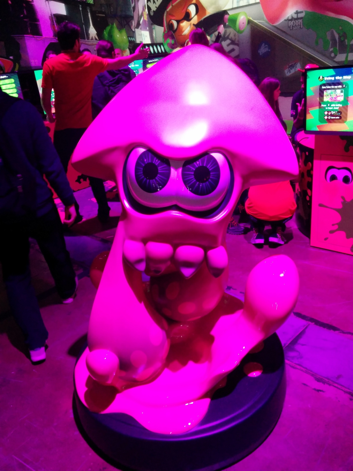Photo of an Inkling statue in the Splatoon 2 area. A giant pink Inkling in squid form is animatedly jumping out of a glossy splash of ink on the statue’s round base, reaching around 165 cm off the ground. Attendees at setups for playing turf wars are visible behind it. Magenta lighting dominates the scene.